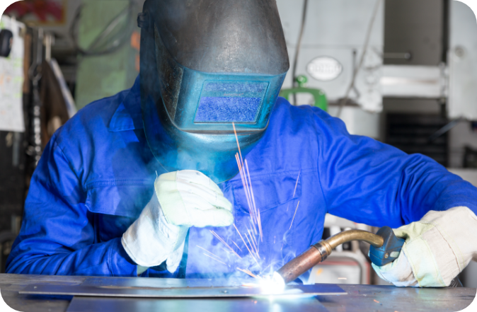 Close up of a welder in gear welding metals on a table. The background workspace is clean and well organized.